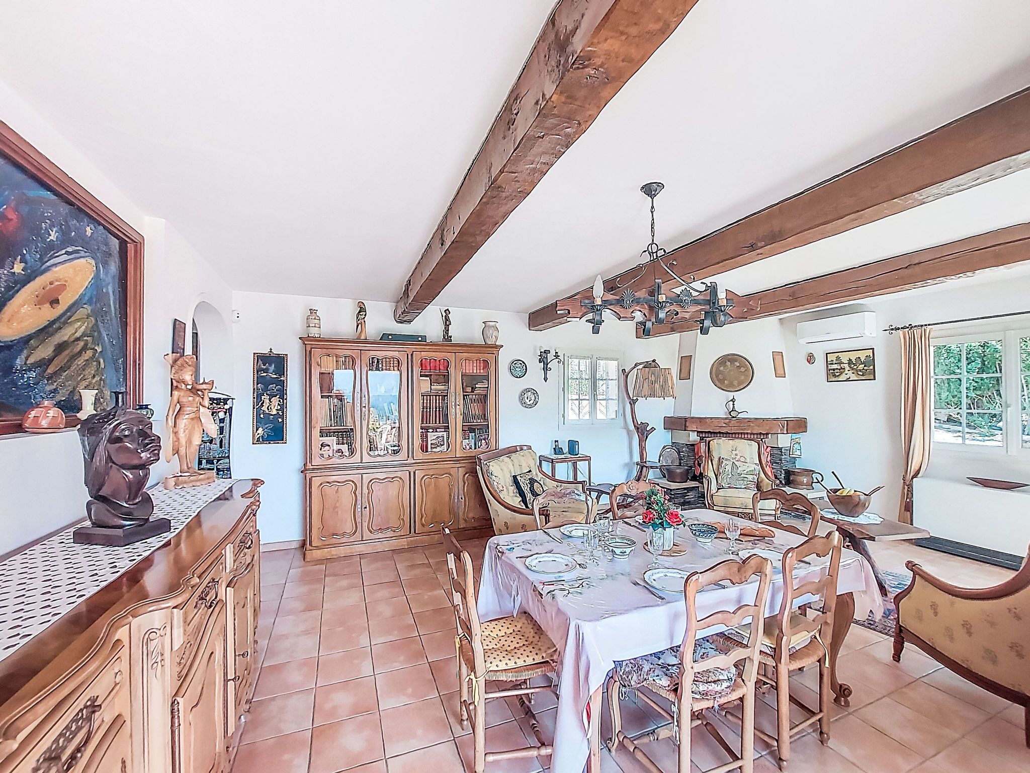 Un coin repas avec une table en bois dressée pour quatre personnes, entourée de chaises, un buffet en bois avec des portes en verre et un grand tableau accroché au mur.