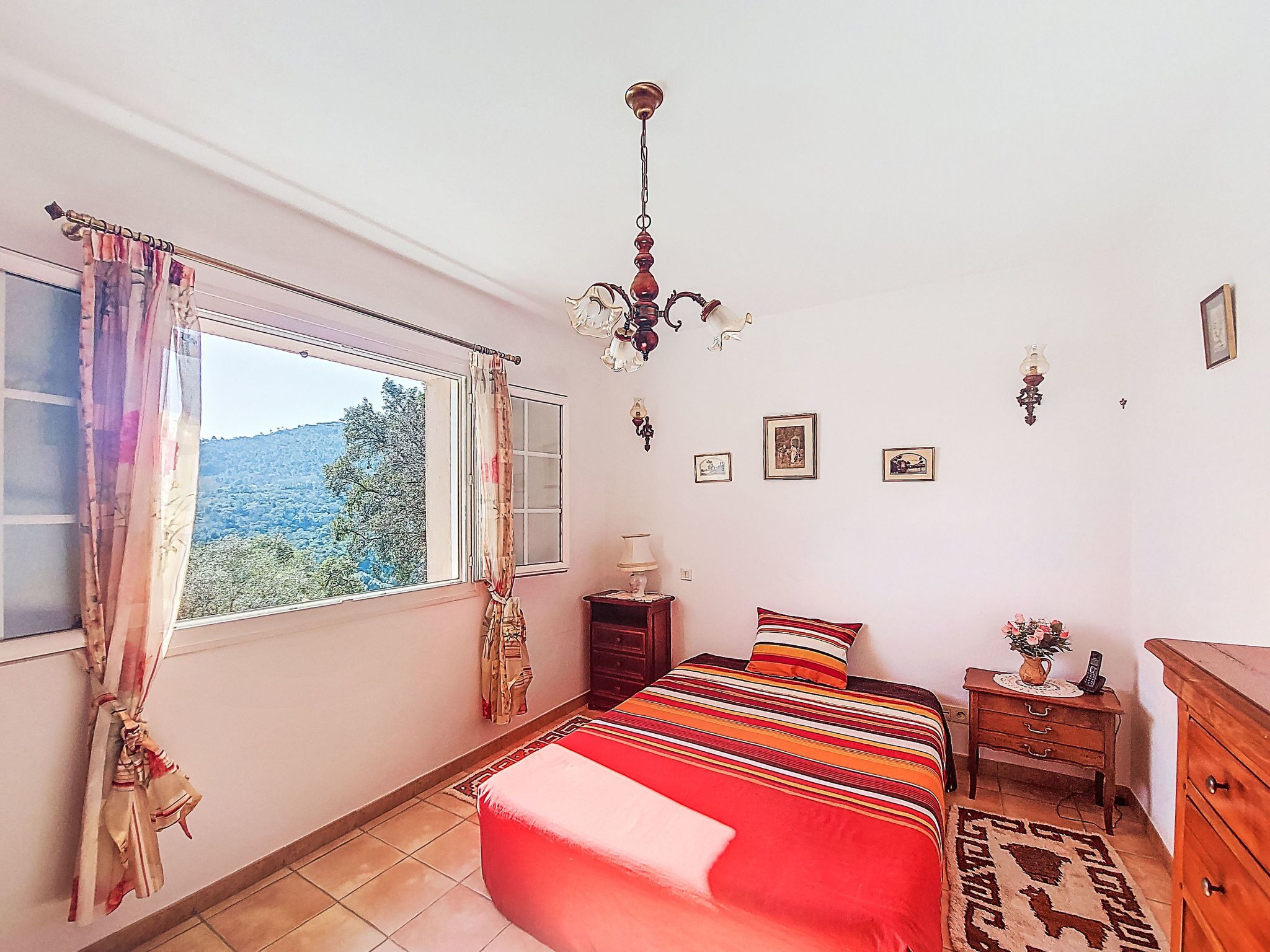 A bedroom with a large window, a bed with a colorful bedspread, a wooden nightstand, a dresser, and a chandelier.
