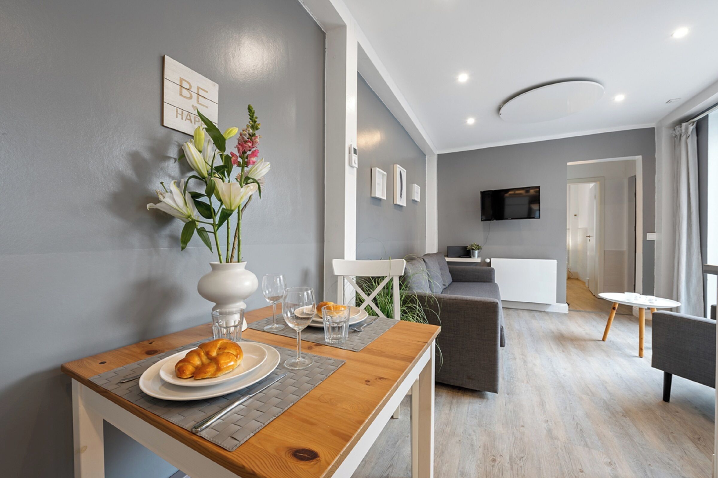 A modern living room with a dining table, a sofa, and a TV mounted on the wall.