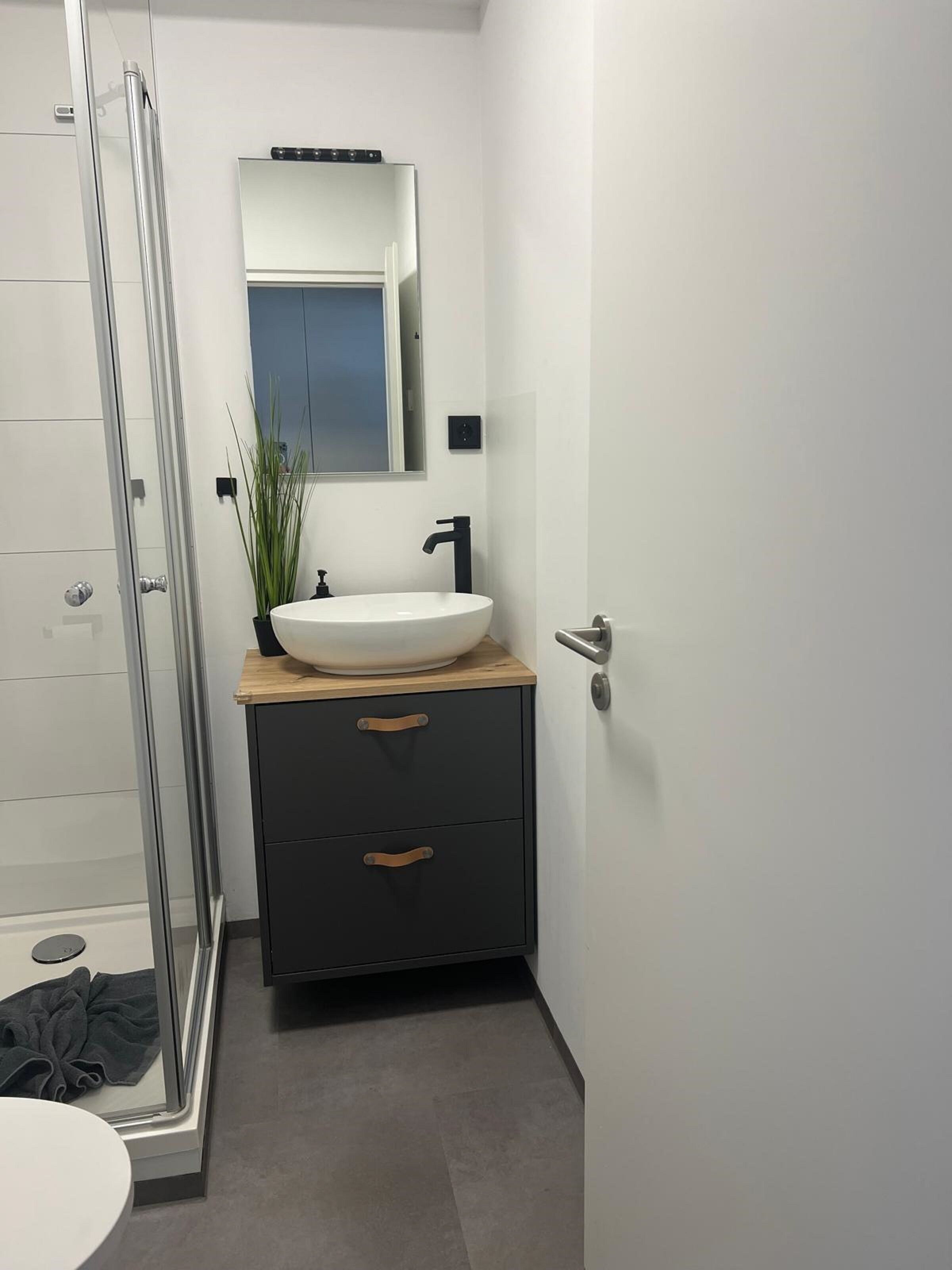 Ein Badezimmer mit Duschkabine aus Glas, einem weißen Waschbecken mit Holztischplatte und einem dunklen Schrank mit Schubladen.