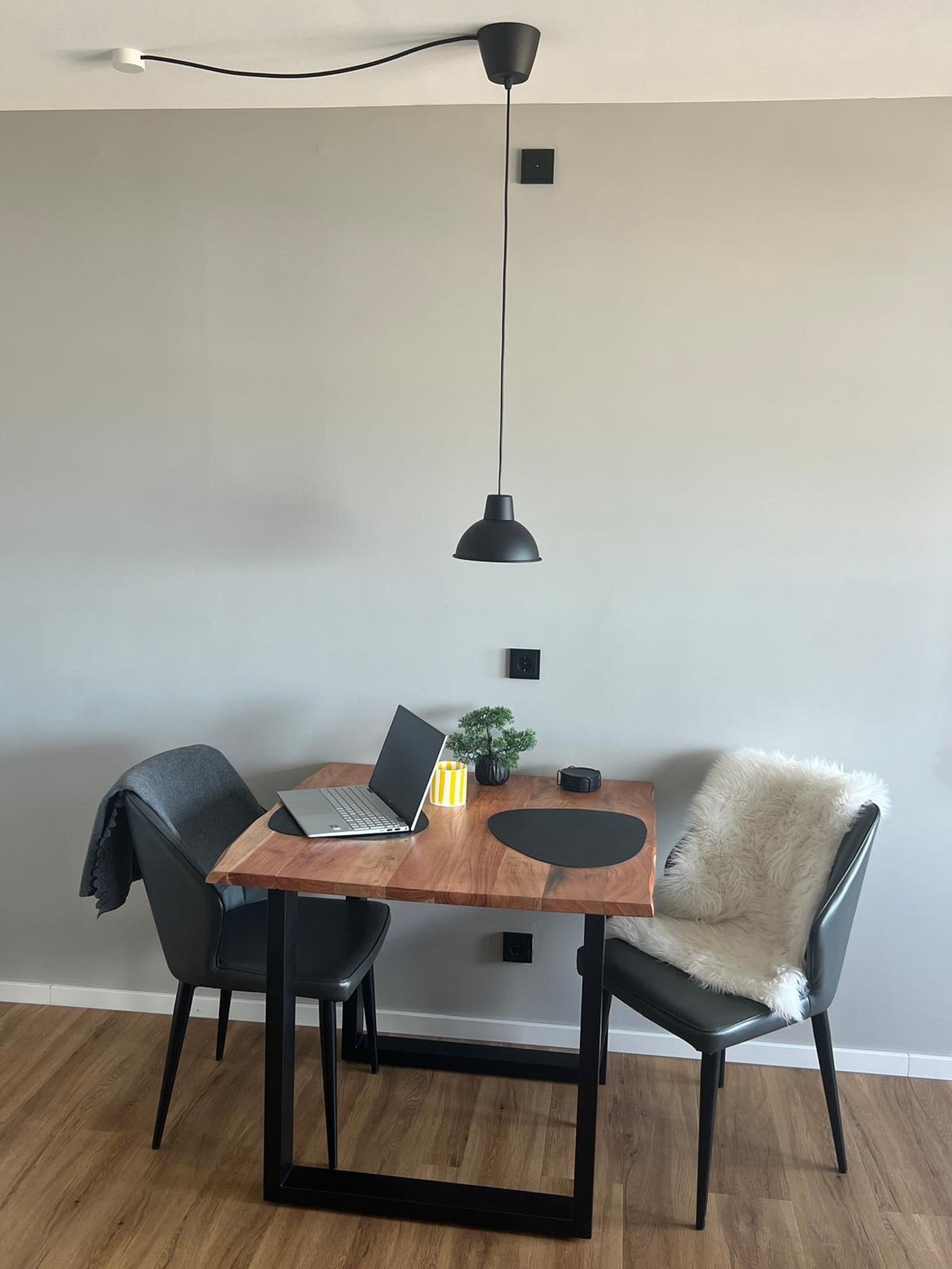 A dining area with a wooden table, two black chairs, a laptop, a small plant, and a black pendant light.