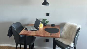 A dining area with a wooden table, two black chairs, a laptop, a small plant, and a black pendant light.