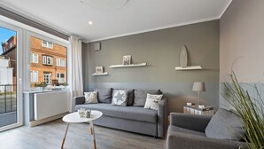 Ein modernes Wohnzimmer mit einem grauen Sofa, einem kleinen runden Tisch und einer Lampe. An der Wand befinden sich Regale mit dekorativen Gegenständen.