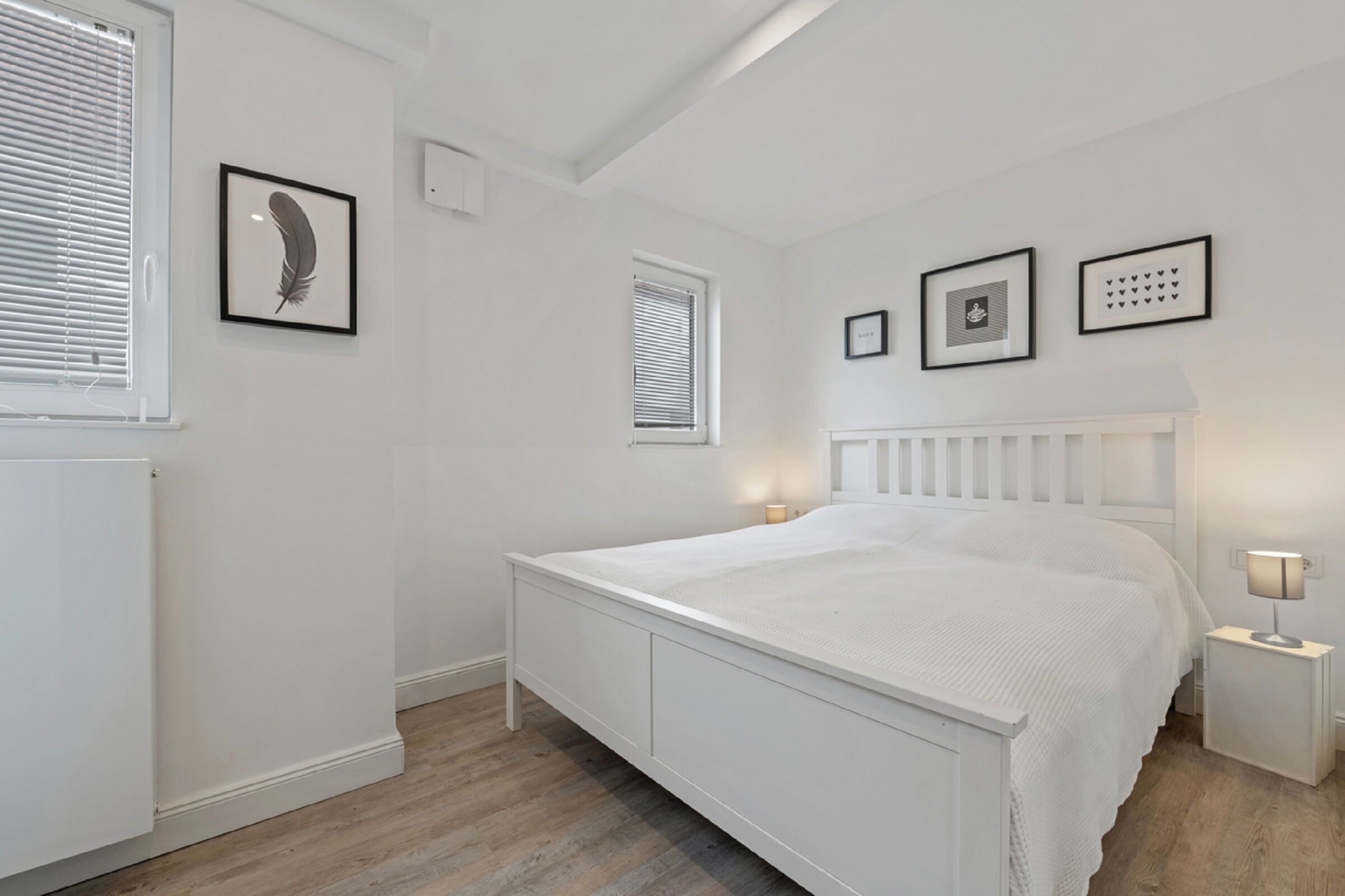 Ein helles Schlafzimmer mit Bett, Nachttischen, einem Heizkörper und gerahmten Bildern an der Wand.