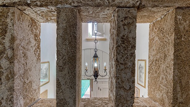 Ein Steinkorridor mit einem Chandelier, gerahmten Bildern an den Wänden und einem Fenster am Ende.
