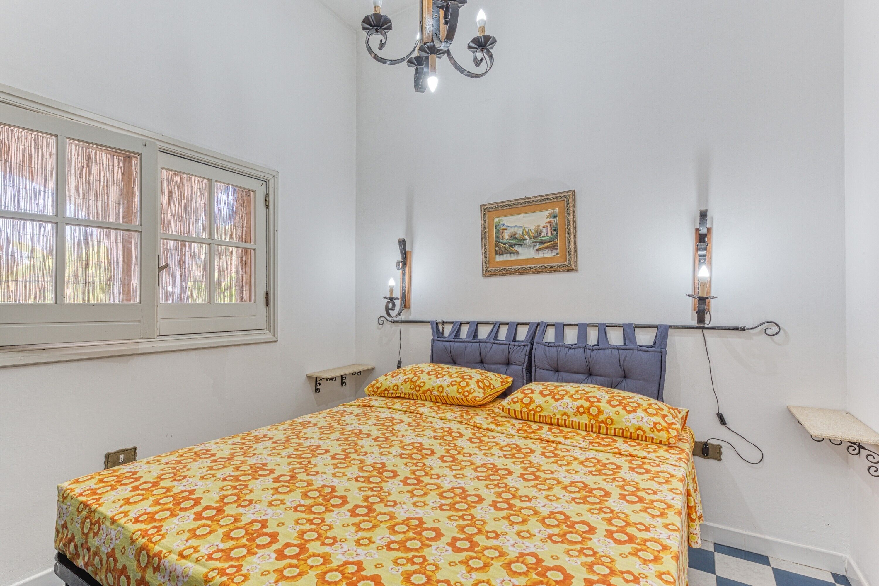 A bedroom with a bed, a window, a chandelier, a wall-mounted picture, and a bedside table.