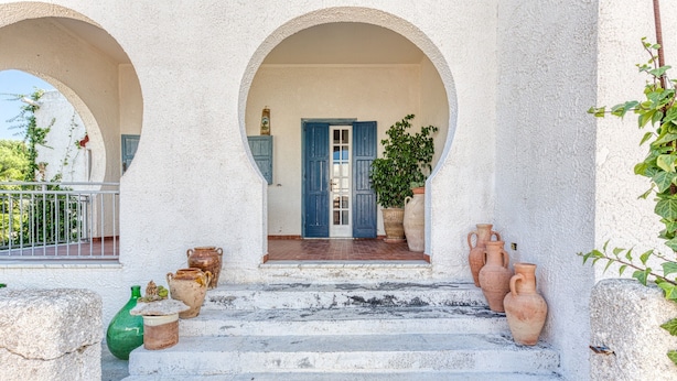 Valkoinen rakennus, jossa on portaikko, joka johtaa siniseen oveen. Rakennuksen ympärillä on ruukuissa olevia kasveja ja terrakottaruukkuja.