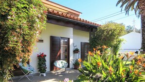 Une maison avec un toit en tuiles, une porte d’entrée et un petit porche agrémenté d’une chaise et de plantes en pot.