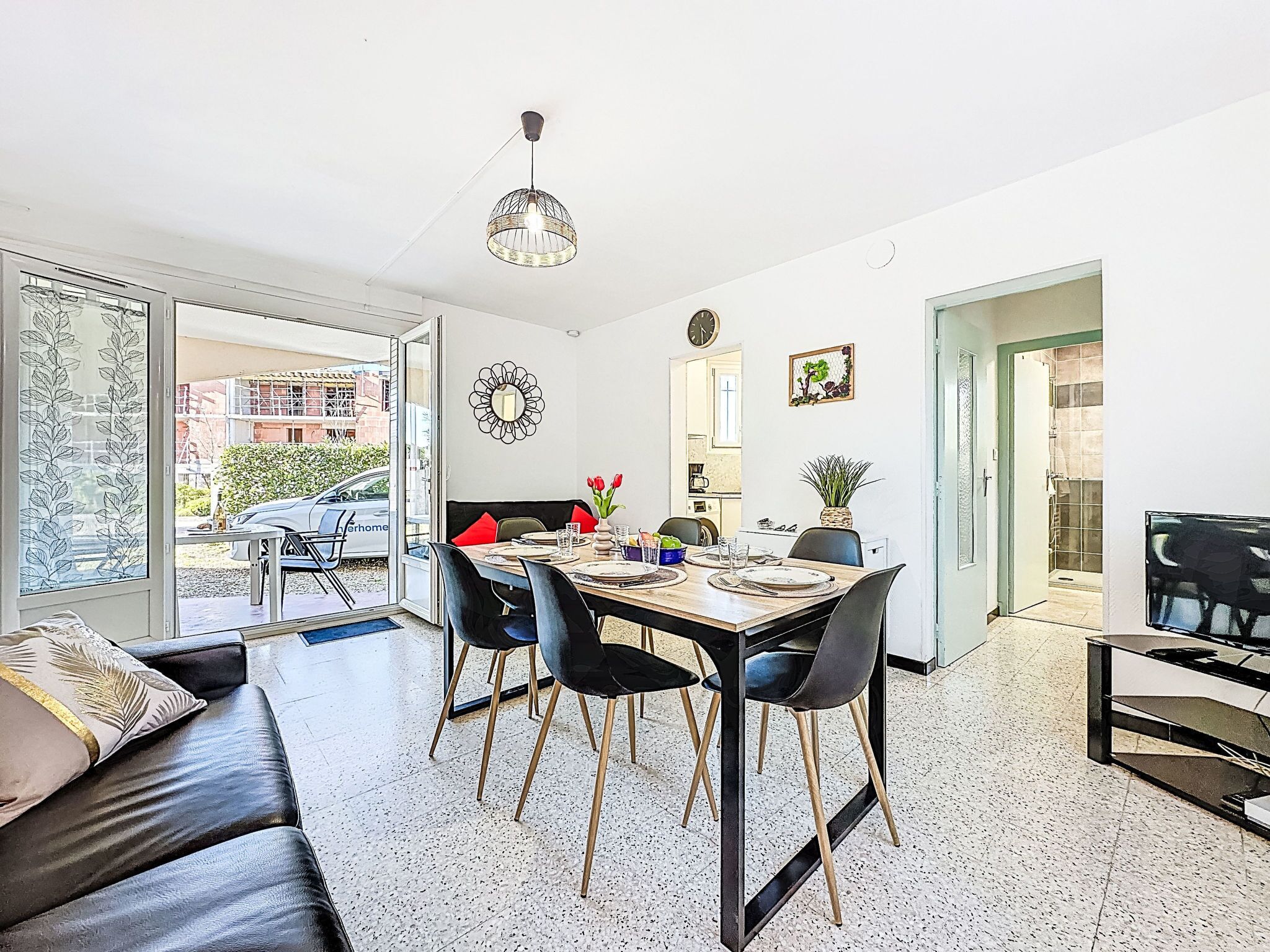 Ein modernes Wohnzimmer mit Essbereich, einer schwarzen Ledersofa, einem Esstisch mit Stühlen, einer Pendelleuchte und Blick ins Freie.