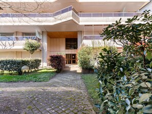 Ein modernes Gebäude mit einem zentralen Eingang, einem überdachten Gang und einem Gartenbereich mit verschiedenen Pflanzen.