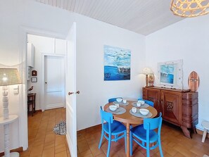Un comedor con mesa de madera y sillas azules, un armario blanco y un cuadro de un paisaje marino.