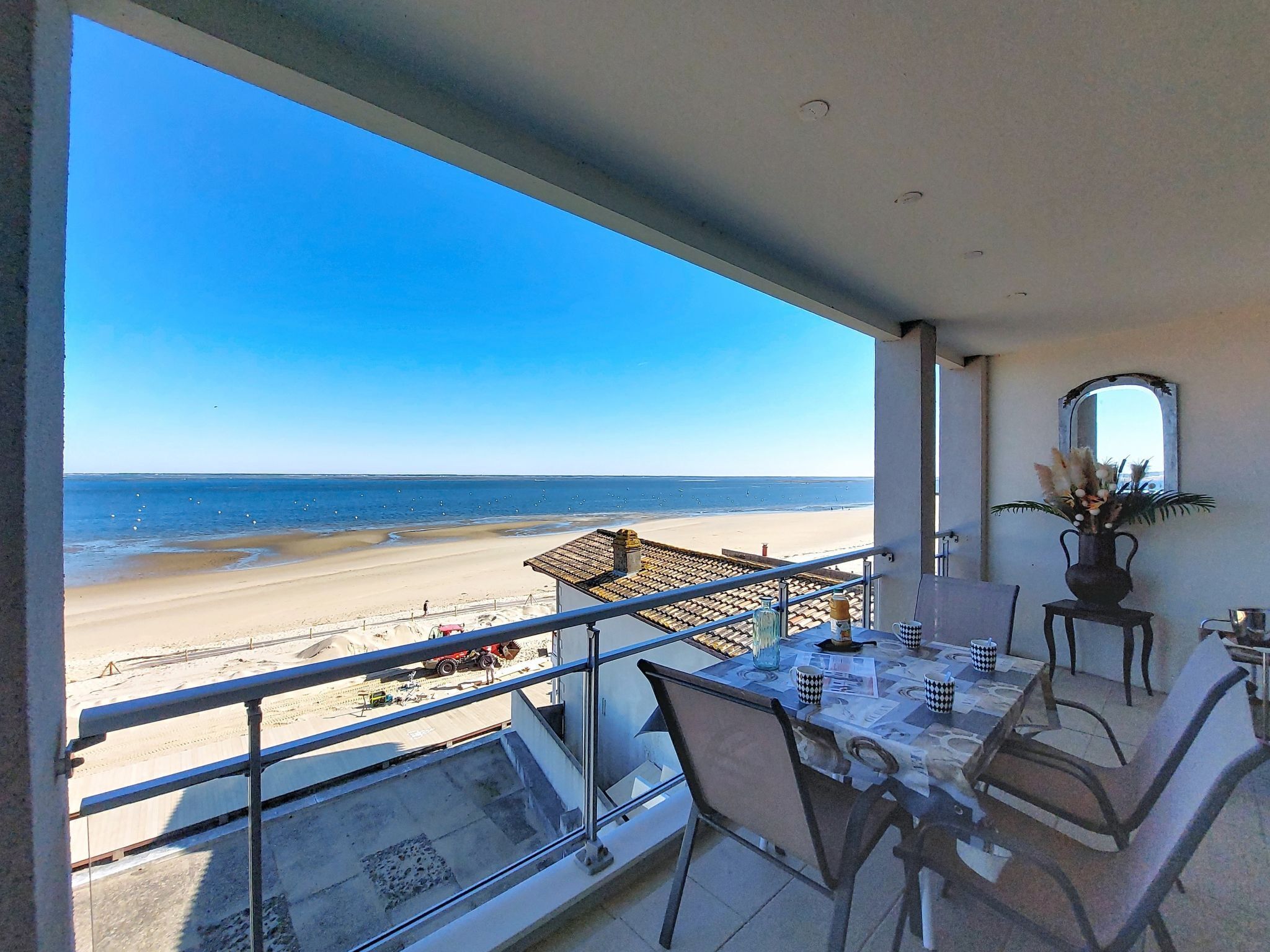 Un balcón con vista a la playa, una mesa de comedor con sillas y un florero con flores.