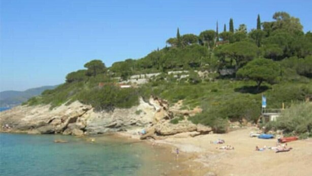 Une plage à l’eau claire, avec un rivage rocheux et des arbres sur une colline.