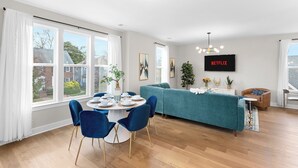 Una sala de estar moderna con un sofá verde azulado, una mesa de comedor con sillas azules y un ventanal con cortinas.