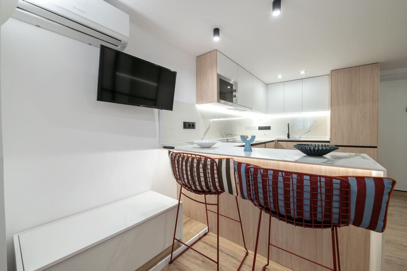 Una cocina moderna con televisor integrado, armarios de madera y una zona de bar con sillas a rayas.