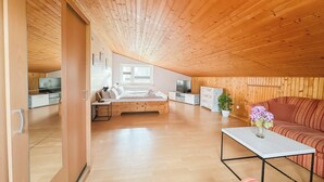 Ein geräumiges Dachzimmer mit geneigter Decke, einem Bett, einer Couch, einem Fernseher und einem Esstisch.