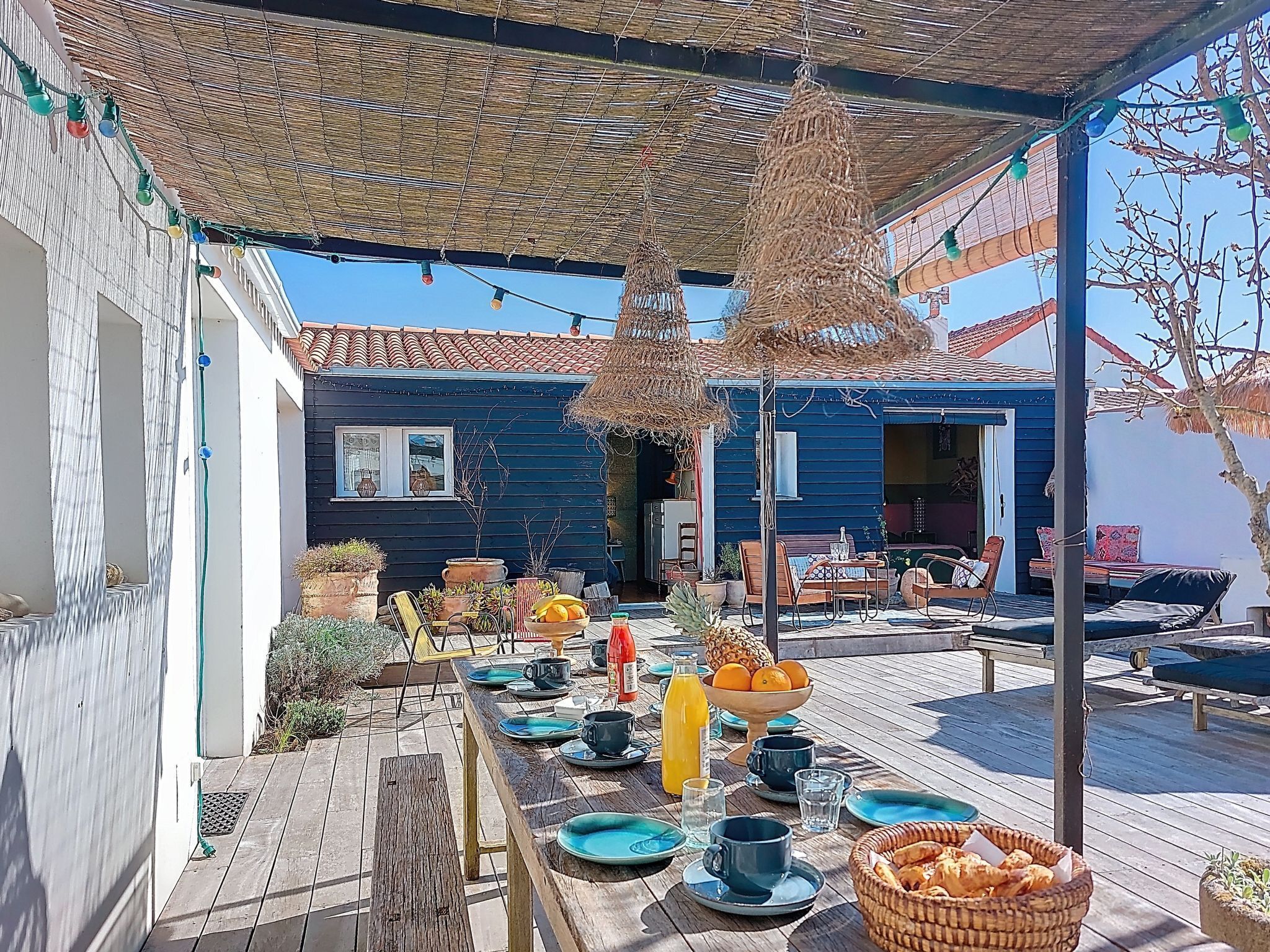 A covered outdoor dining area with a table set for breakfast, a blue house in the background, and a thatched roof overhead.