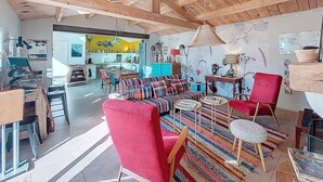 Een ruime woonkamer met een hoog houten plafond, een kleurrijk gestreept tapijt, een rode fauteuil, en een eethoek in de achtergrond.