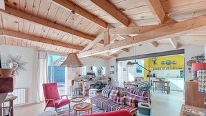 Een ruime woonkamer met een hoog houten plafond, een kleurrijke bank, een rode fauteuil en een eethoek die door een open doorgang zichtbaar is.