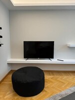 Una sala de estar moderna con un televisor de pantalla plana sobre un estante blanco, un reposapiés negro y un suelo de madera con estampado tipo espiga.