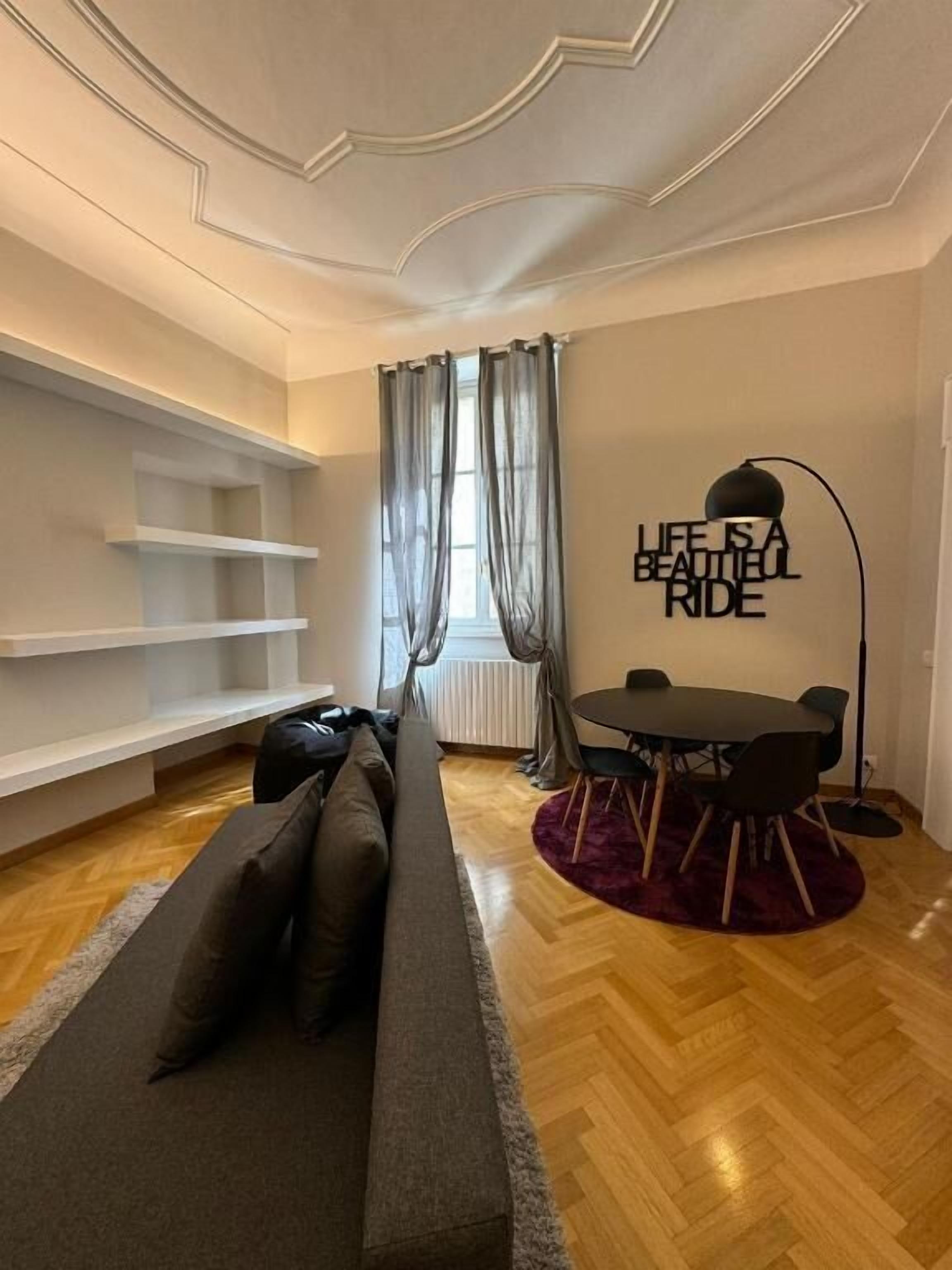 Een moderne woonkamer met een bank, een eettafel met stoelen, een vloerlamp en een raam met dunne gordijnen.