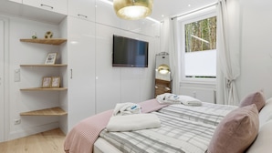 A bedroom with a bed, a TV mounted on the wall, built-in shelves, a window with curtains, and a ceiling light fixture. - Vacation apartment "Waldduft" between the forest and the Baltic Sea beach (Heringsdorf)
