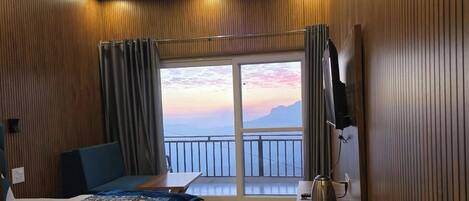 Habitación de hotel con cama, escritorio, silla, televisor y balcón con vistas a las montañas, además de una ventana con cortinas.