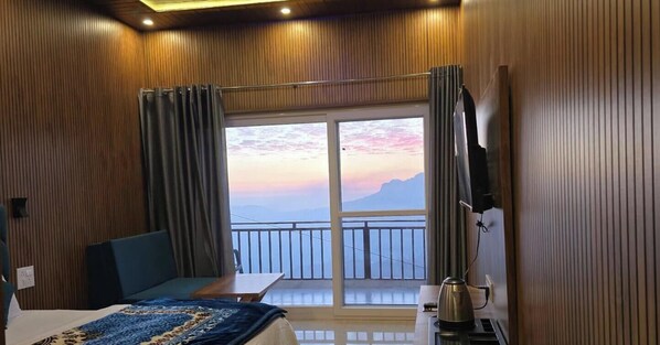 A hotel room with a bed, a desk, a chair, a TV, a balcony with a view of mountains, and a window with curtains. - Milan Home Stay (Tehri)