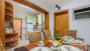 Un coin repas avec une table en bois dressée pour quatre personnes, des chaises en osier et une cuisine en arrière-plan.