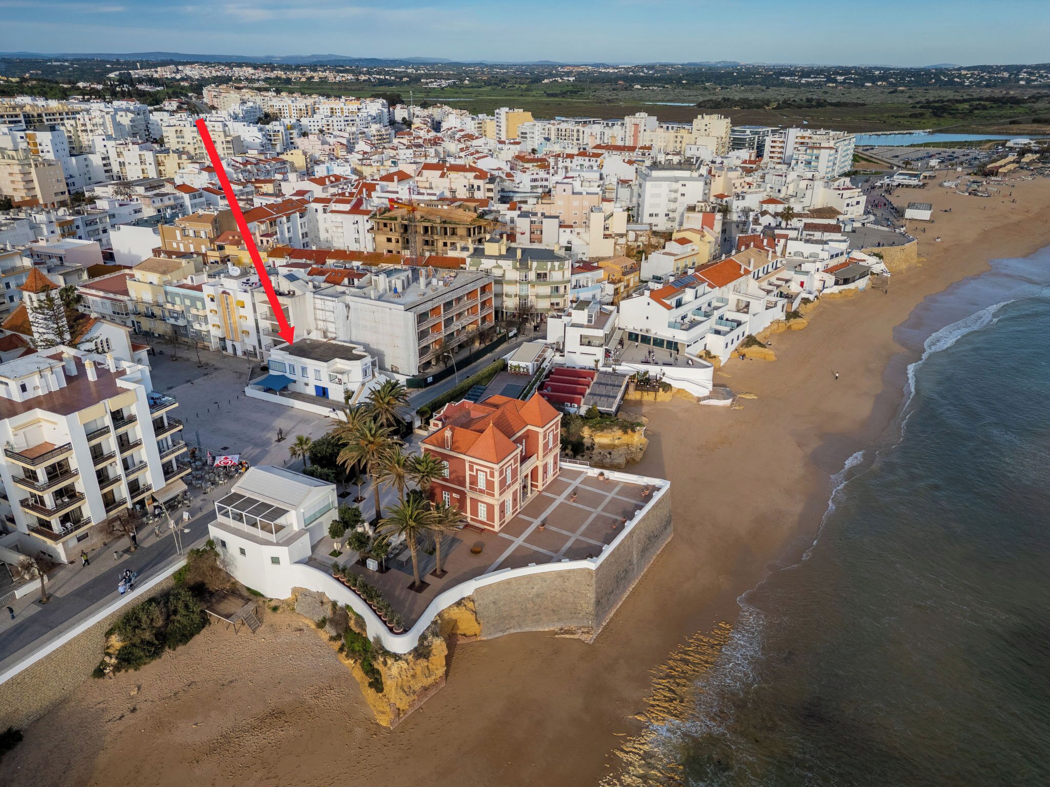 Luftaufnahme einer Küstenstadt mit Gebäuden, einem Strand und einem roten Pfeil, der auf einen bestimmten Ort zeigt.