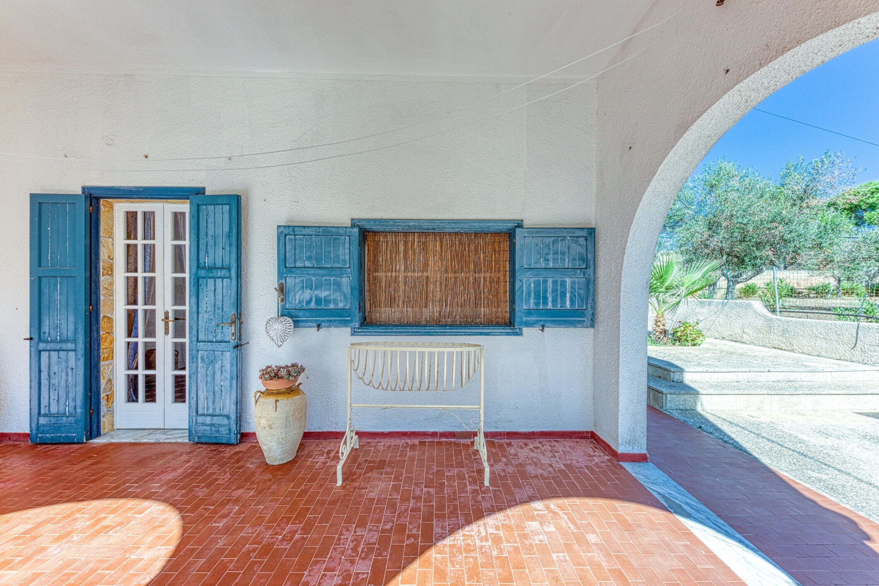 Une pièce avec un sol carrelé, une porte blanche avec des volets bleus, un radiateur blanc et une vue sur la verdure à l’extérieur.