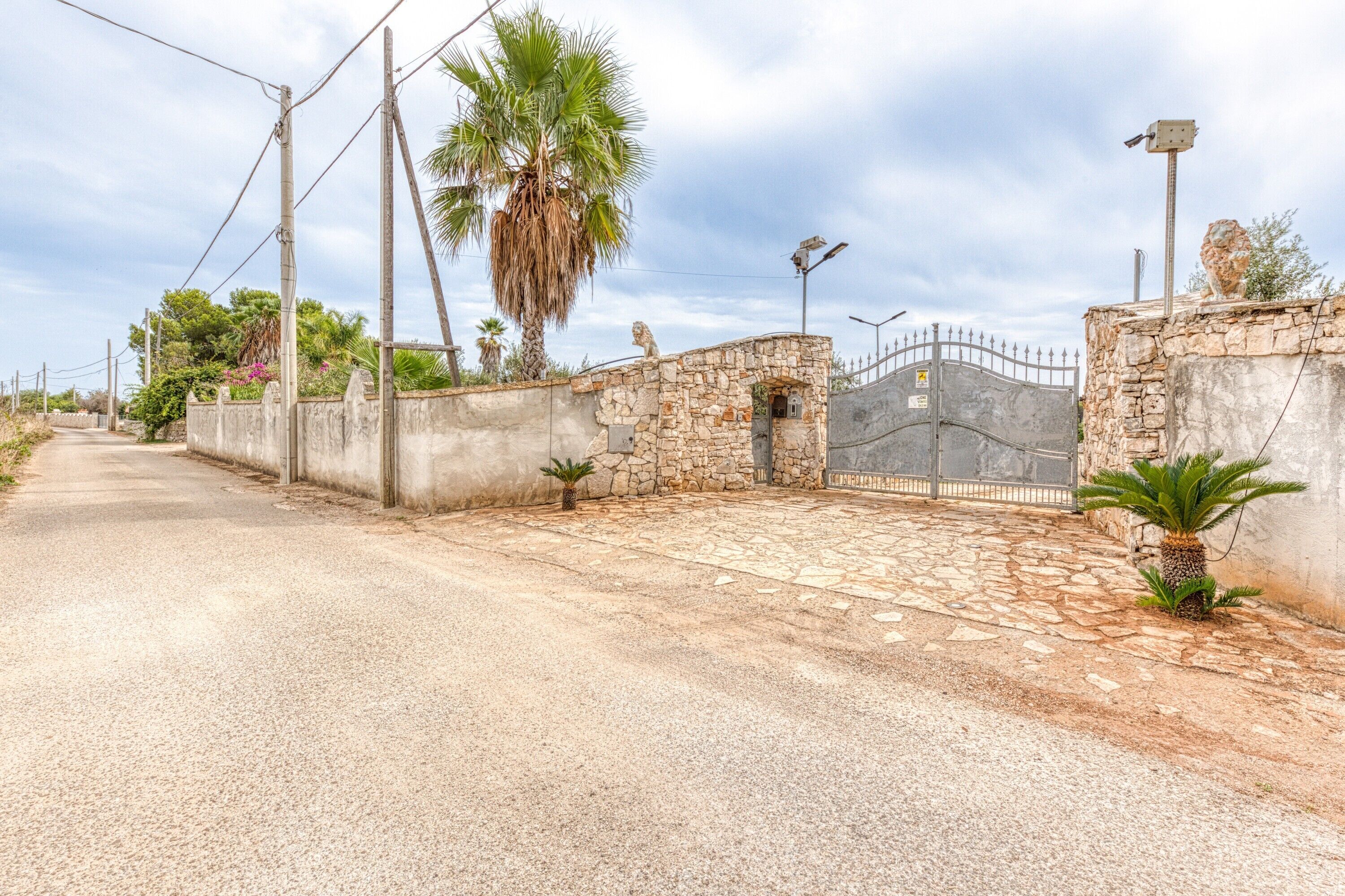 Une route asphaltée mène à une entrée fermée par un portail, avec un mur en pierre, un palmier et un portail en métal.