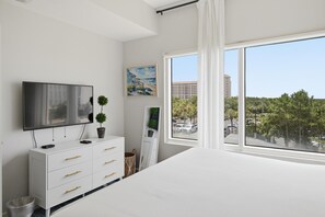 Ein Schlafzimmer mit großem Fenster, einem Flachbildfernseher, einer weißen Kommode und einem Bild an der Wand.