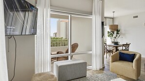 Una sala de estar con televisor, sofá, mesa de comedor con sillas y balcón con vista a edificios.