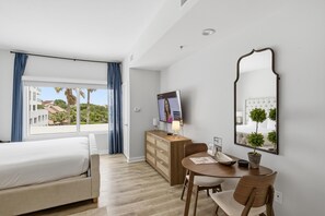 Habitación de hotel con cama, escritorio y silla, televisor, cómoda y un amplio ventanal con cortinas.