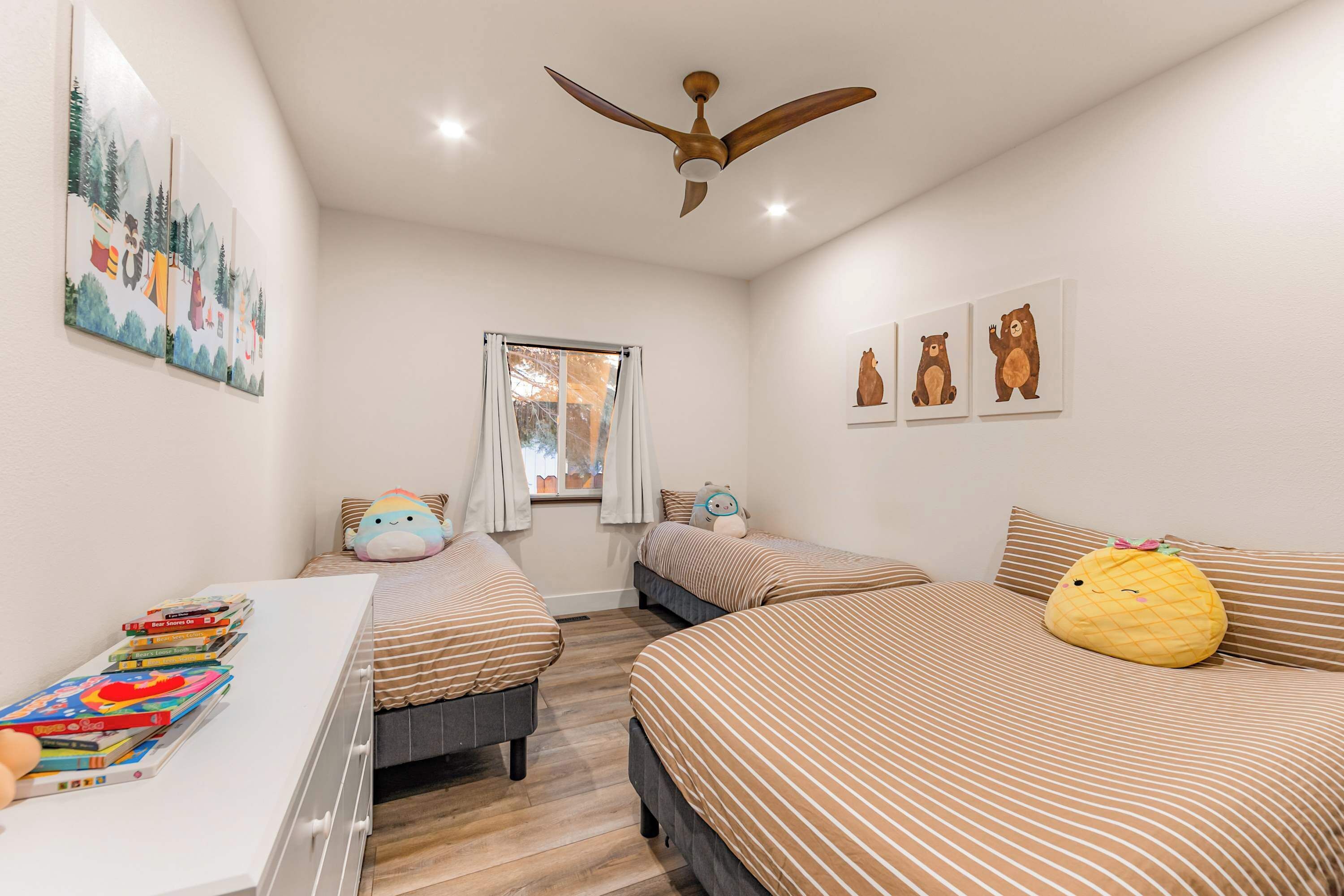 A bedroom with two beds, a ceiling fan, a window with curtains, wall art, and a bedside table with books.