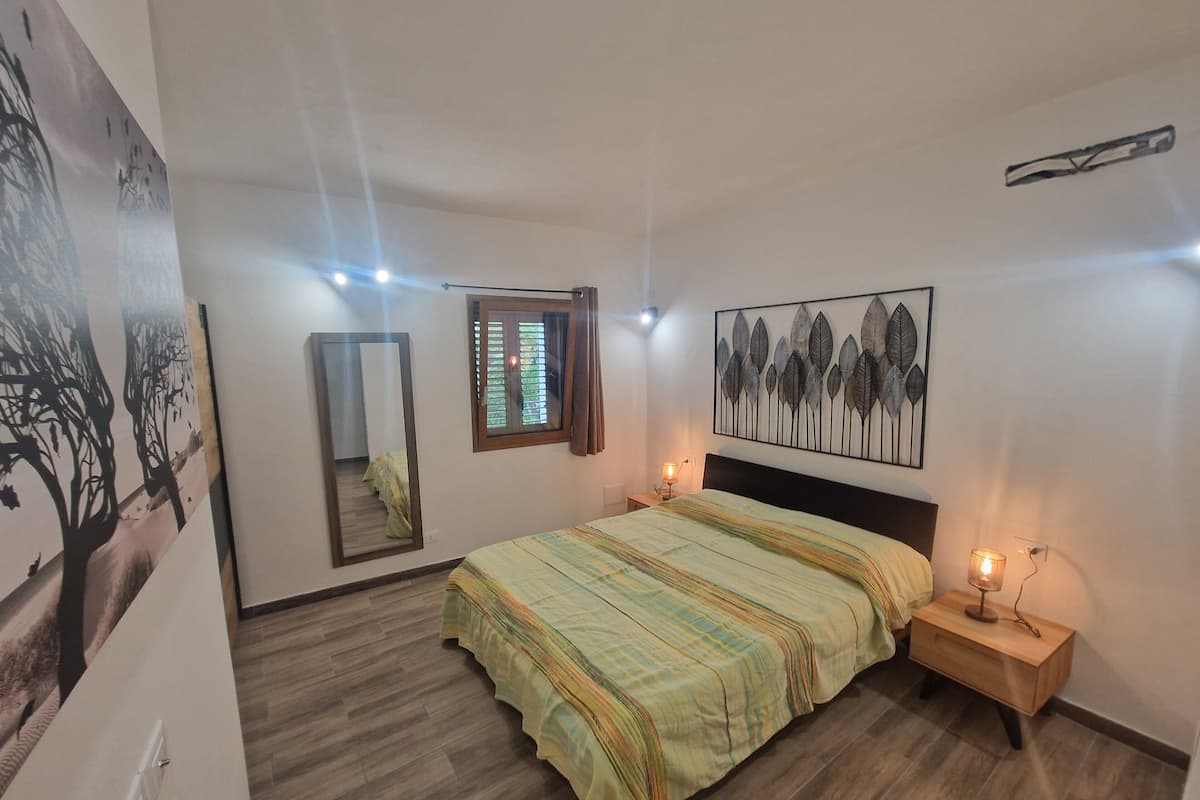 A bedroom with a bed, a nightstand, a wall-mounted mirror, a window with blinds, and a wall art piece.