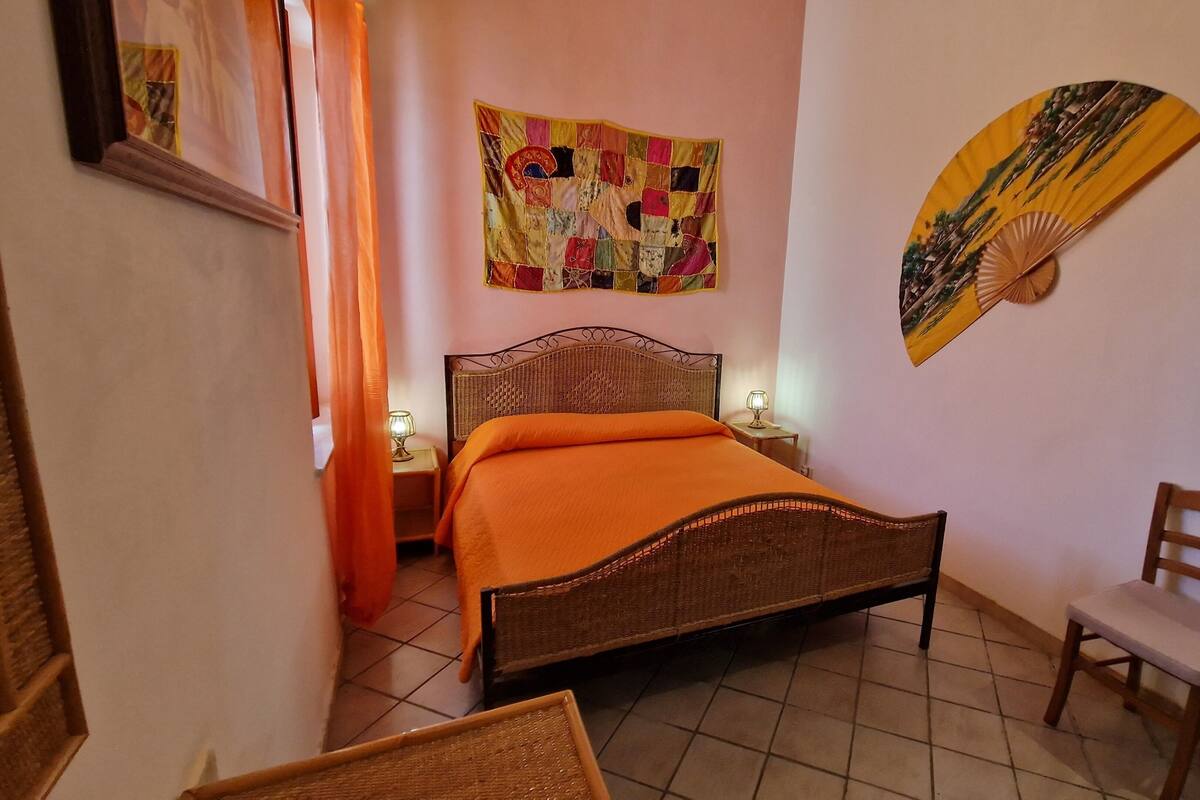 A narrow room with a bed, a chair, a wall tapestry, and a wall fan.