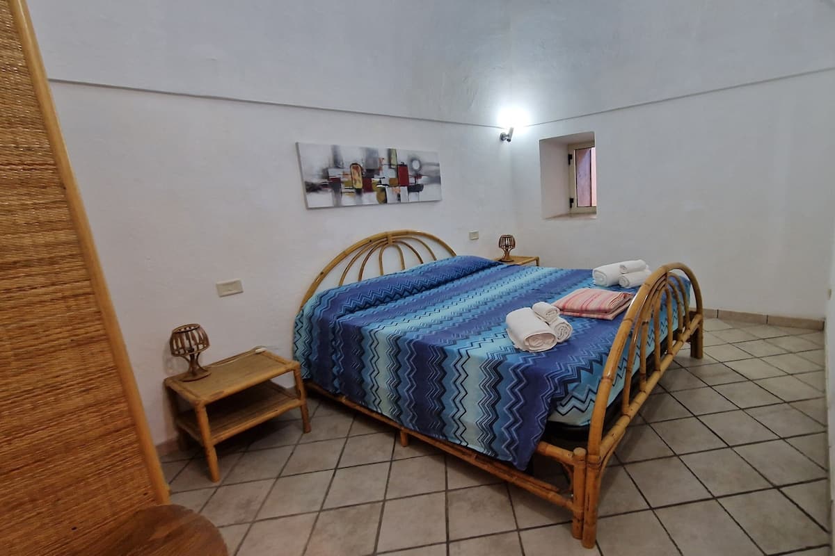 A bedroom with a bed, a small bedside table, a wall-mounted light, and a window.