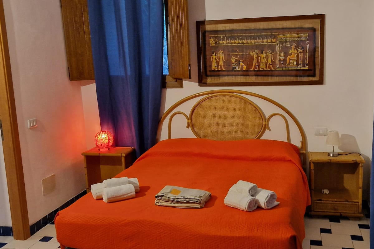 A hotel room with a bed, a nightstand with a lamp, a wall-mounted painting, and a window with curtains.