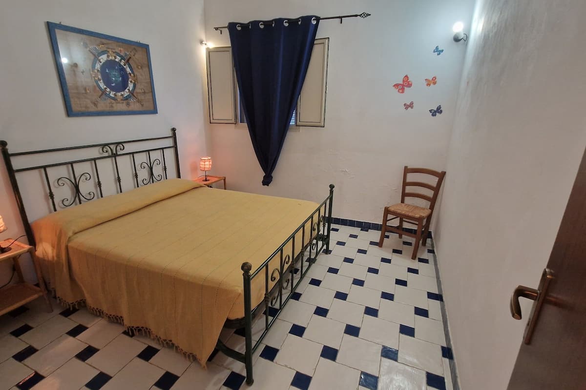 A bedroom with a bed, a chair, a nightstand, a lamp, a curtain, a wall clock, and a decorative wall piece.