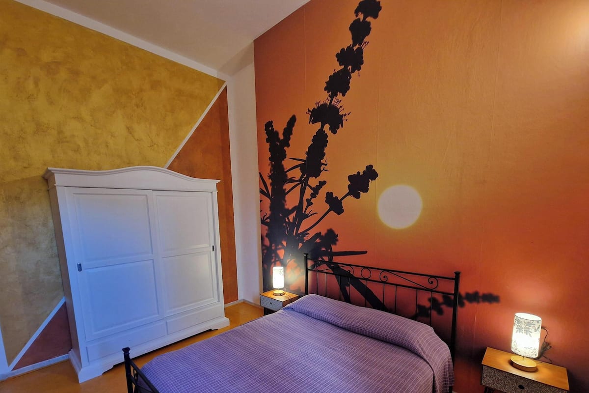 A bedroom with a bed, a nightstand with a lamp, a wardrobe, and a wall with a floral mural.