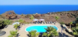 A pool with lounge chairs and umbrellas, surrounded by palm trees and a view of the ocean.