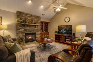 Una sala de estar con chimenea de piedra, televisor, muebles de cuero y ventilador de techo.