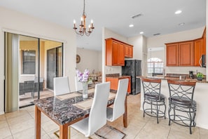 A modern kitchen with granite countertops, wooden cabinets, a black refrigerator, and a dining area with a marble table and white chairs. - Charmz Retreat - with shared pool (Kissimmee)