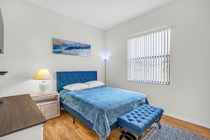 Un dormitorio con cama, lámpara de noche, banco, obra de arte, persianas y alfombra.