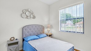 Un dormitorio con cama, mesita de noche, lámpara, ventana con persianas y un adorno en la pared.