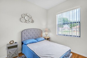 A bedroom with a bed, a bedside table, a lamp, a window with blinds, and a wall decoration.