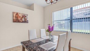 Comedor con una mesa de mármol, sillas blancas, un jarrón con flores púrpuras, un cuadro en la pared y una ventana con persianas.