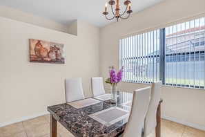 Comedor con una mesa de mármol, sillas blancas, un jarrón con flores púrpuras, un cuadro en la pared y una ventana con persianas.